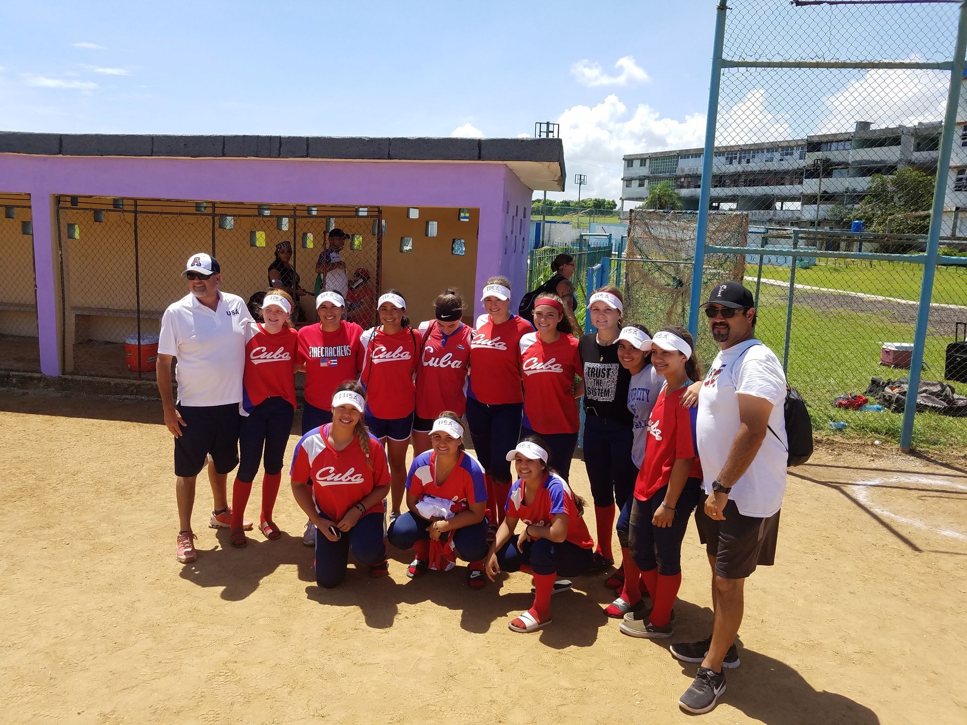 Softball team in red jerseys posing on a dirt field. Coaches standing at the sides.