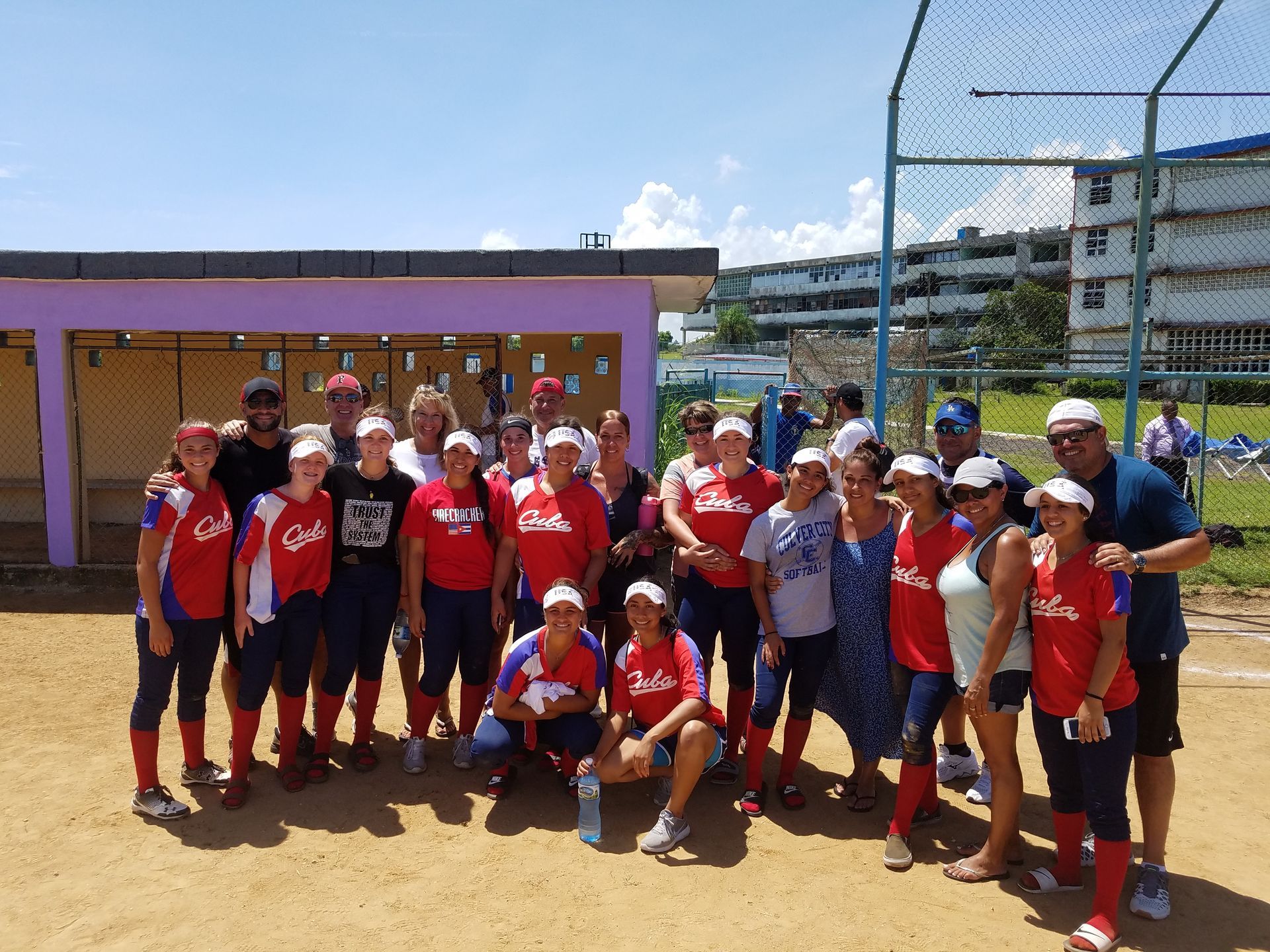 A group of people wearing red shirts pose on a dirt field. A dugout and buildings are visible in the background.
