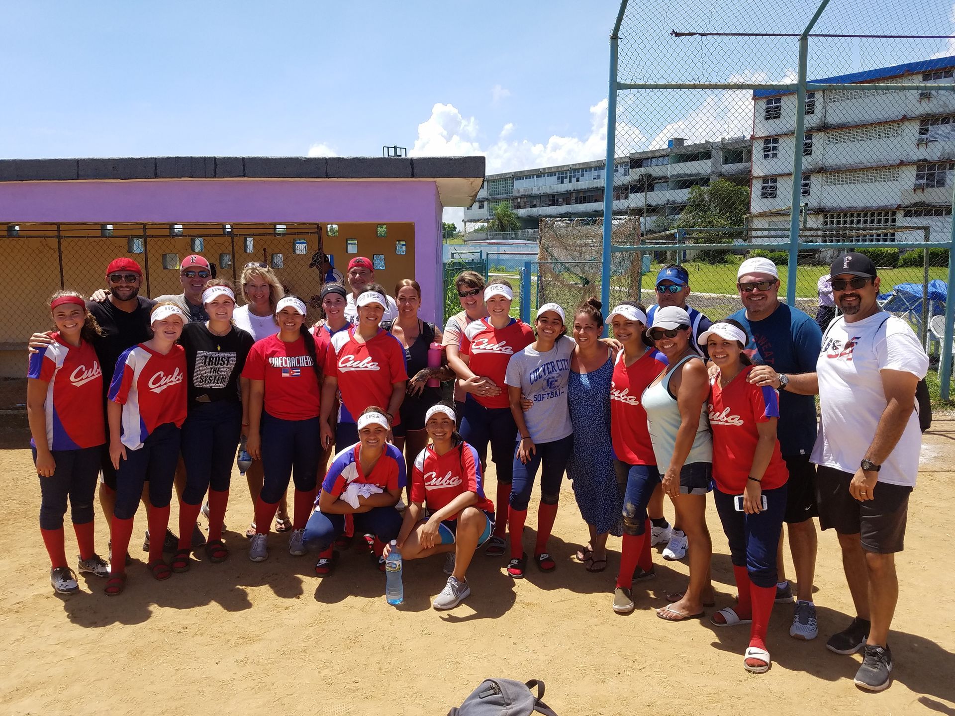 A softball team in red and blue uniforms poses on a sandy field, near a building and fence on a sunny day.