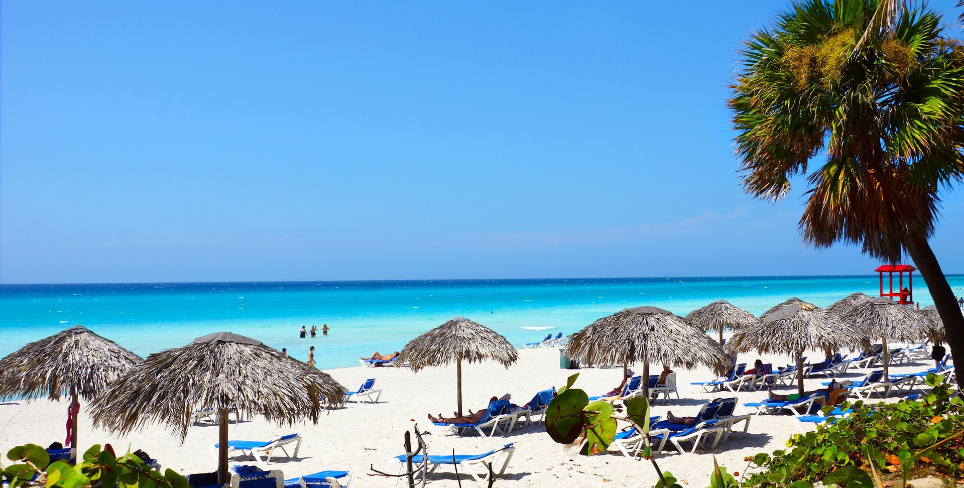Beach scene with white sand, turquoise water, thatched umbrellas, and palm trees under a blue sky.