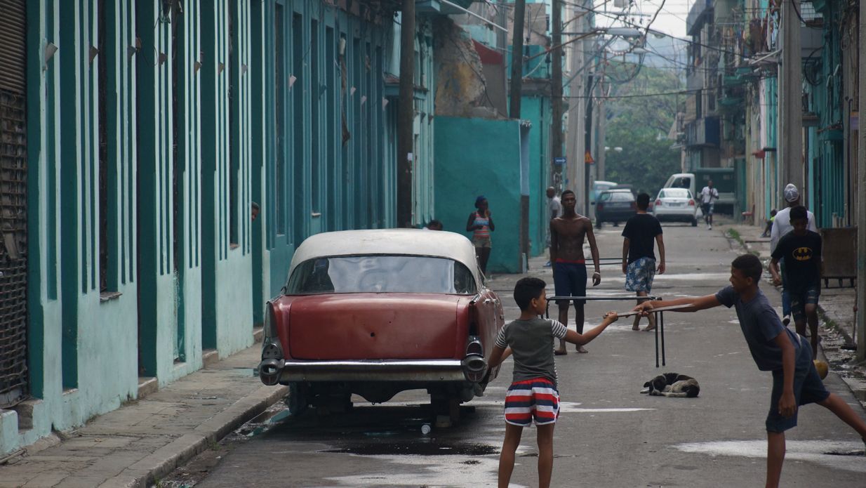 Children play in street with vintage car in Havana, Cuba. Teal buildings line the street.
