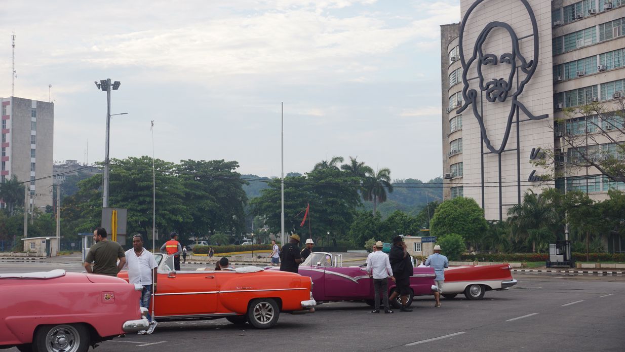Classic cars parked on a Havana street with people nearby; a large mural is on the building in the background.