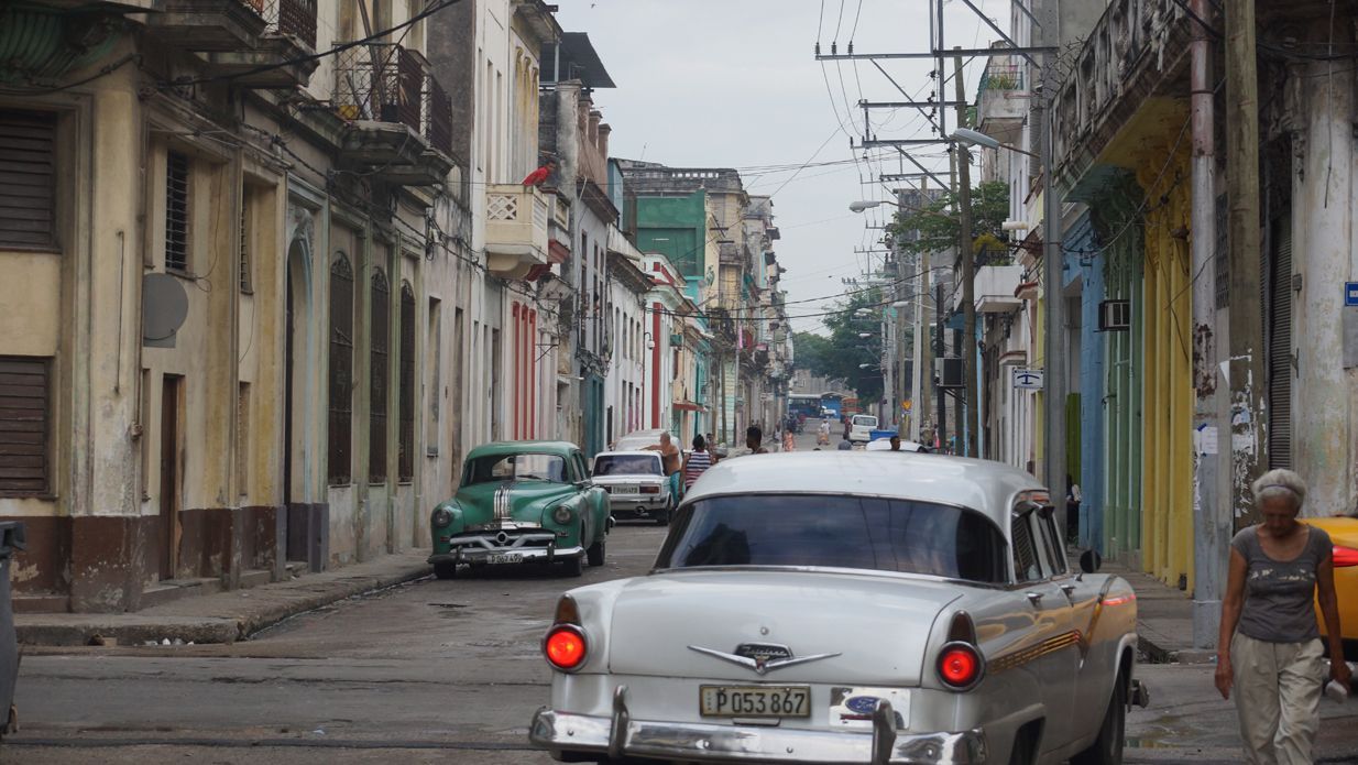Classic cars travel down a street in Havana, Cuba, lined with colorful buildings.