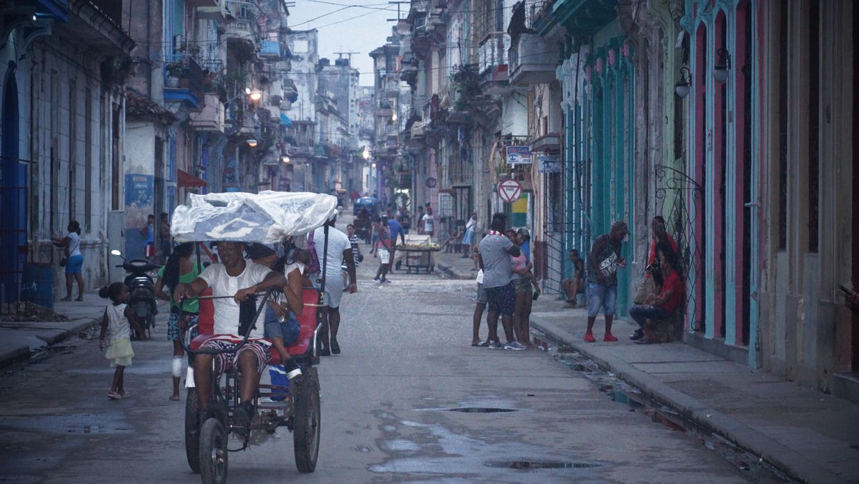 Street scene in Havana, Cuba, with people, buildings with colorful facades, and a three-wheeled vehicle carrying a large load.