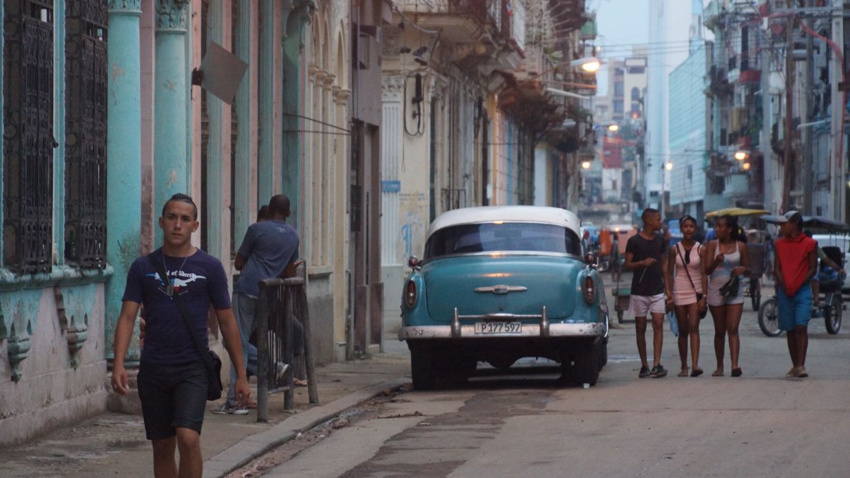 Street scene in Havana, Cuba, with classic turquoise car parked. People walking, pastel buildings, dusk lighting.