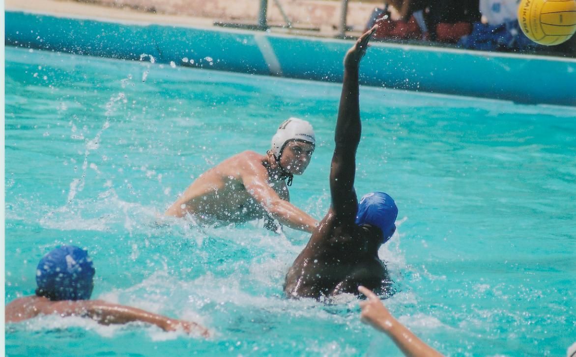 Water polo players in a pool. One raises an arm as others attempt to block. Bright blue water.