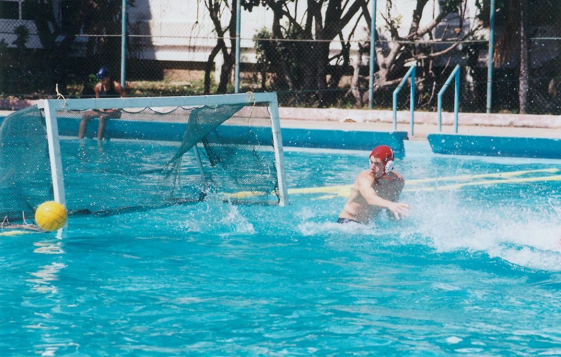 Water polo player in red cap in pool, goalie defends the net. Yellow ball near the goal.