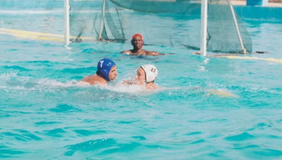 Water polo players in a pool, one with a blue cap and one with a white cap, goalie watches the action.