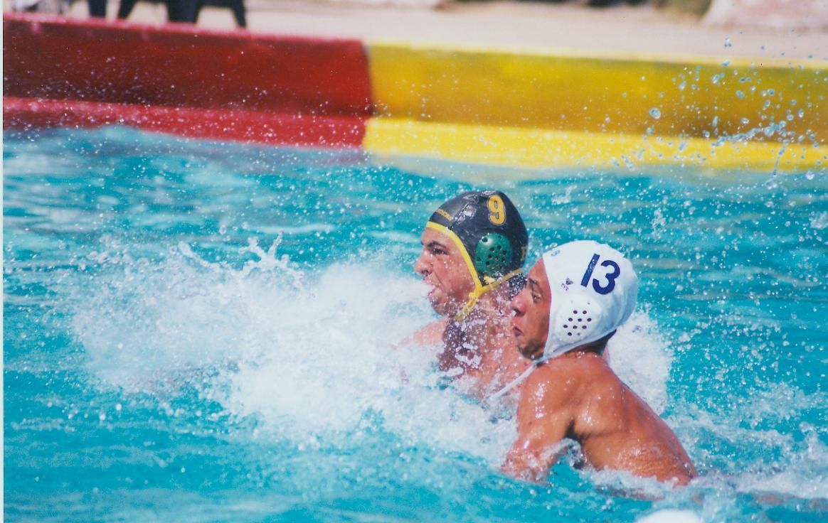 Water polo players in action, one in green cap, one in white with number 13, splashing in pool.