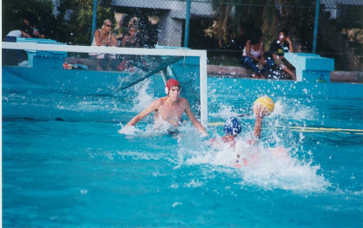 Water polo game: player in white with ball about to shoot; goalie in red cap defends.