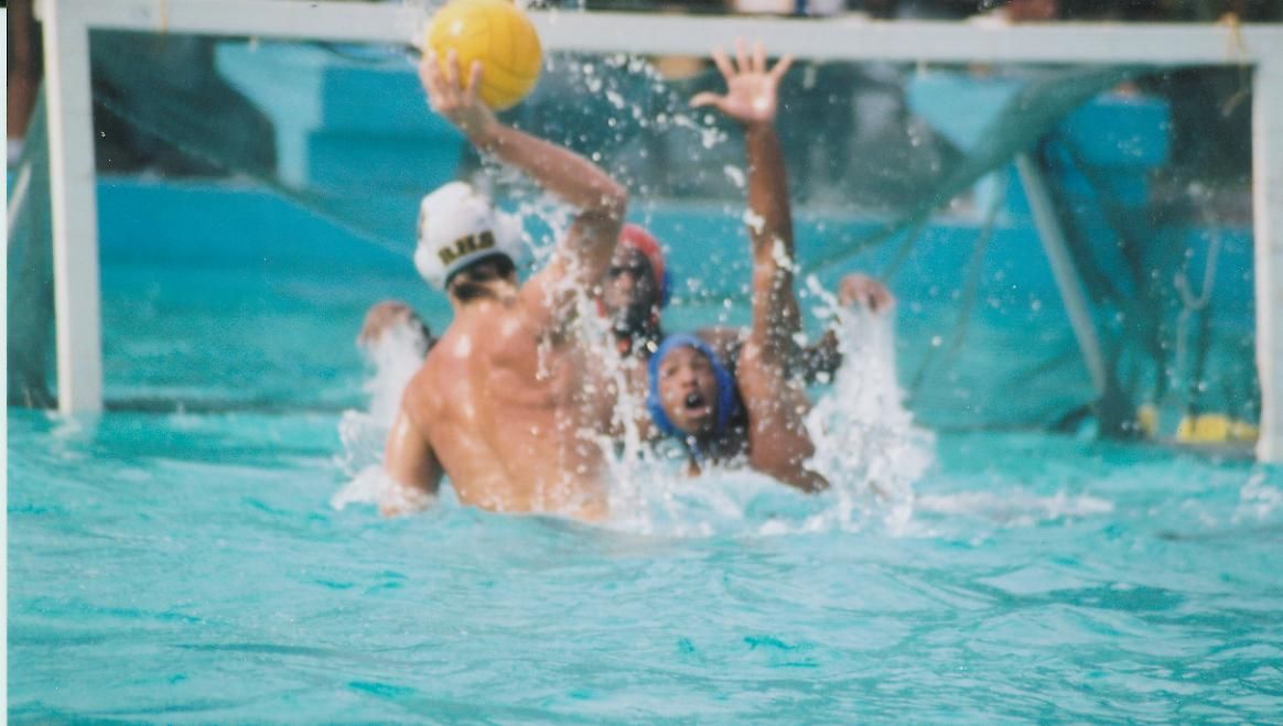 Water polo players in a pool: one throws ball, two others attempt to block the goal.