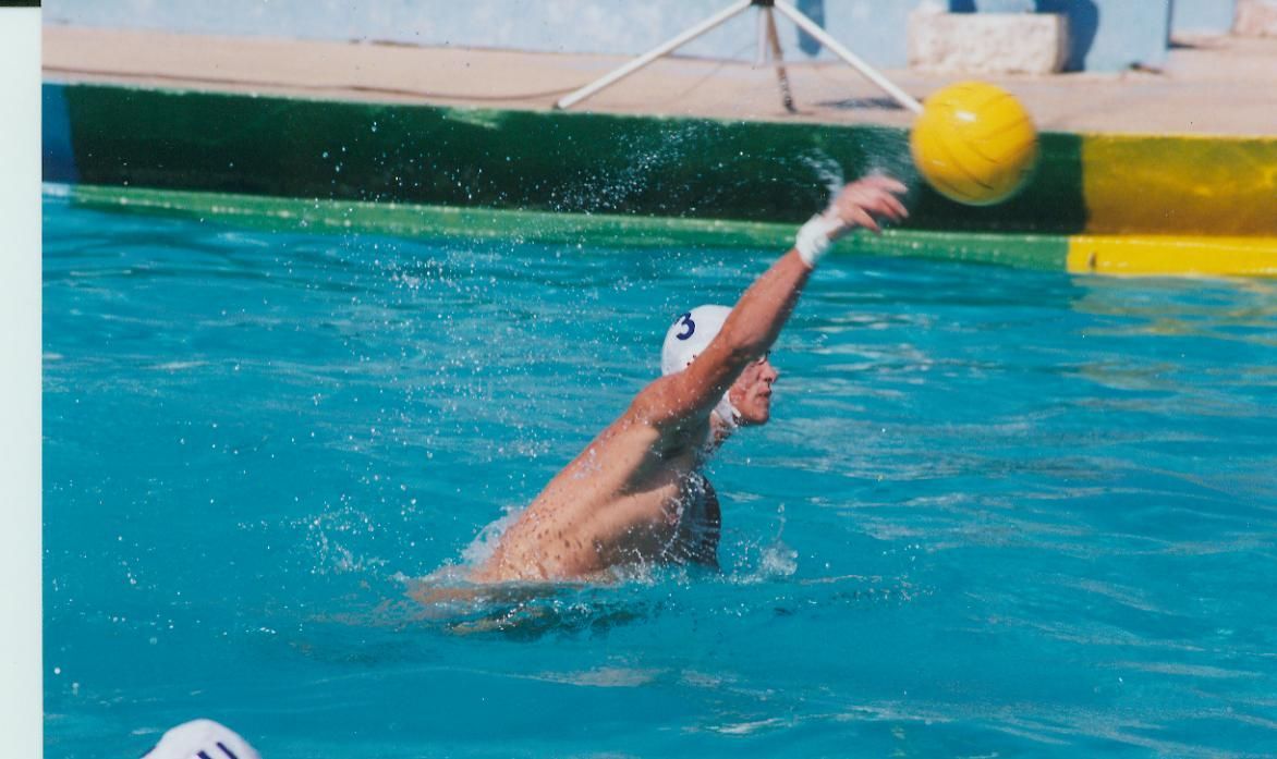 Water polo player throwing ball in pool, wearing white cap.