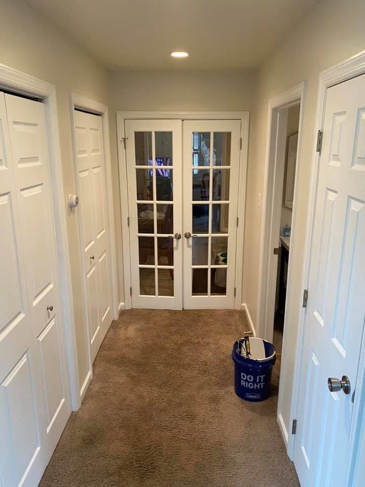 Narrow carpeted hallway with white doors and glass French doors at the end, blue bucket on the floor.