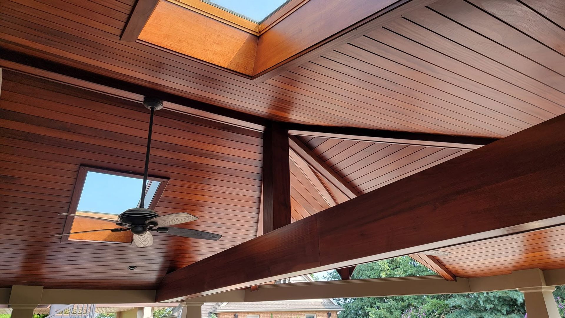 Covered wooden patio ceiling with a ceiling fan and exposed beams, lit by warm daylight.