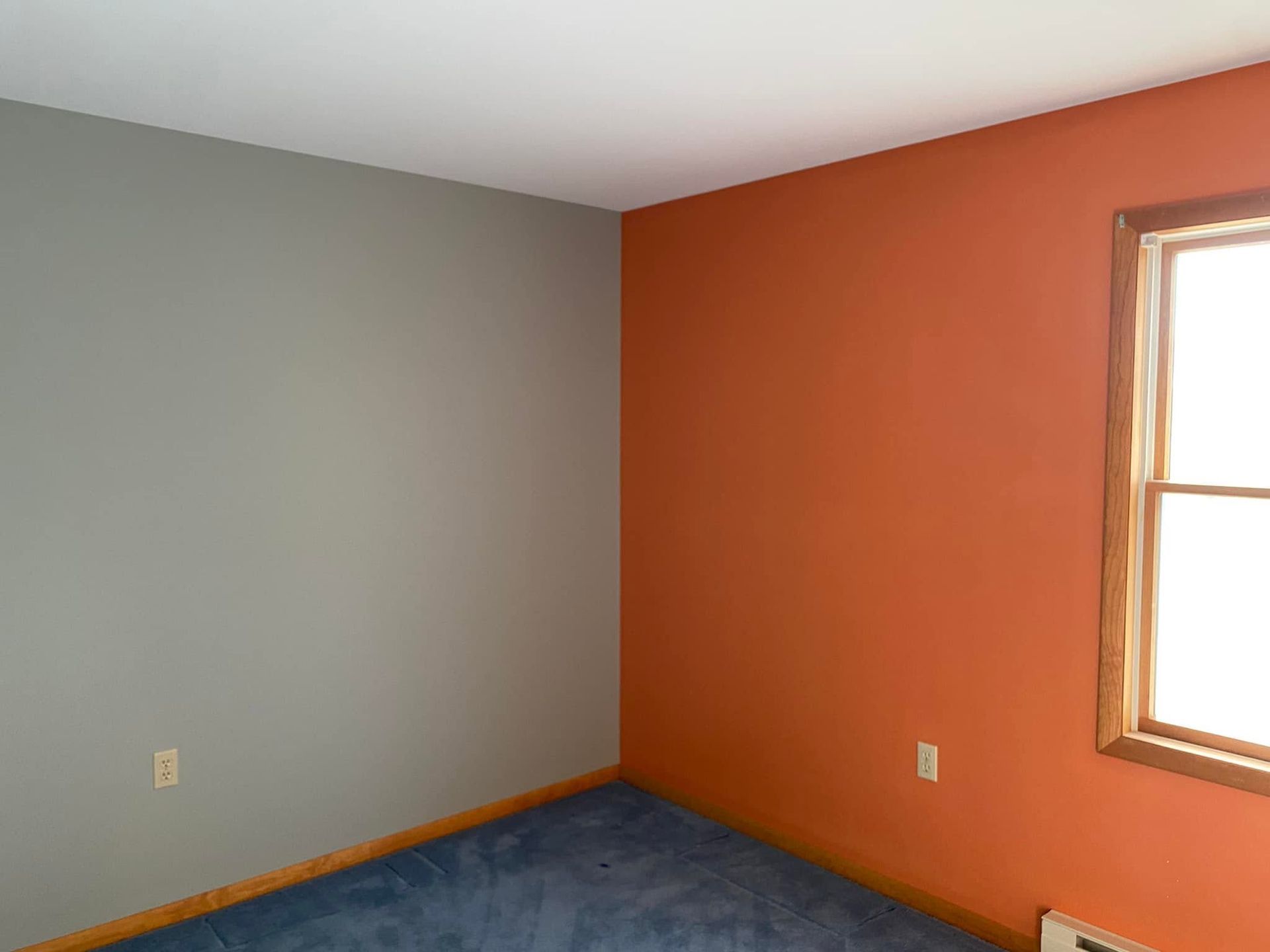 Empty room with orange and gray walls, blue carpet, and a window letting in daylight.