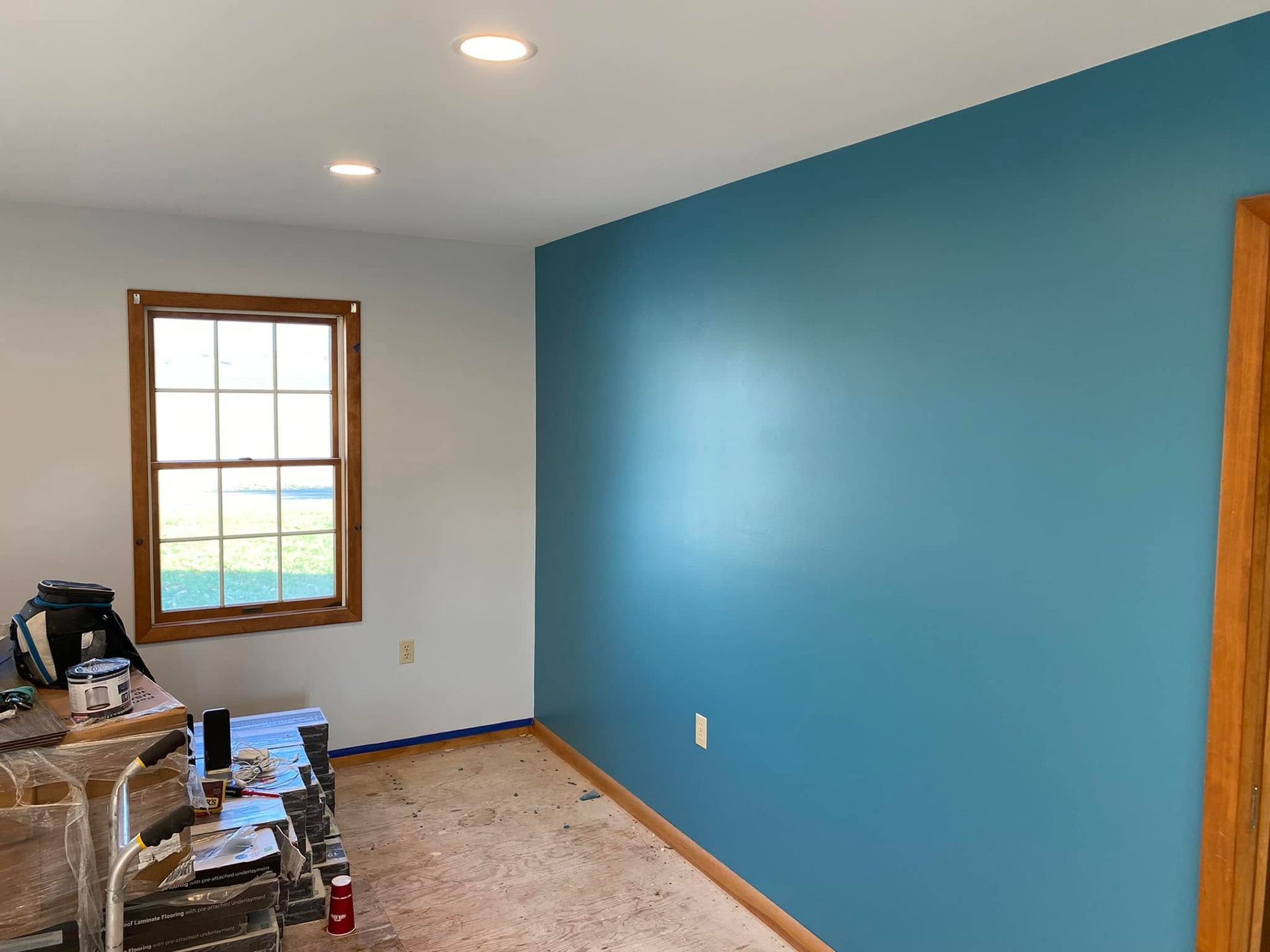 Blue accent wall in an empty room with a window, ceiling lights, and construction items on the floor.