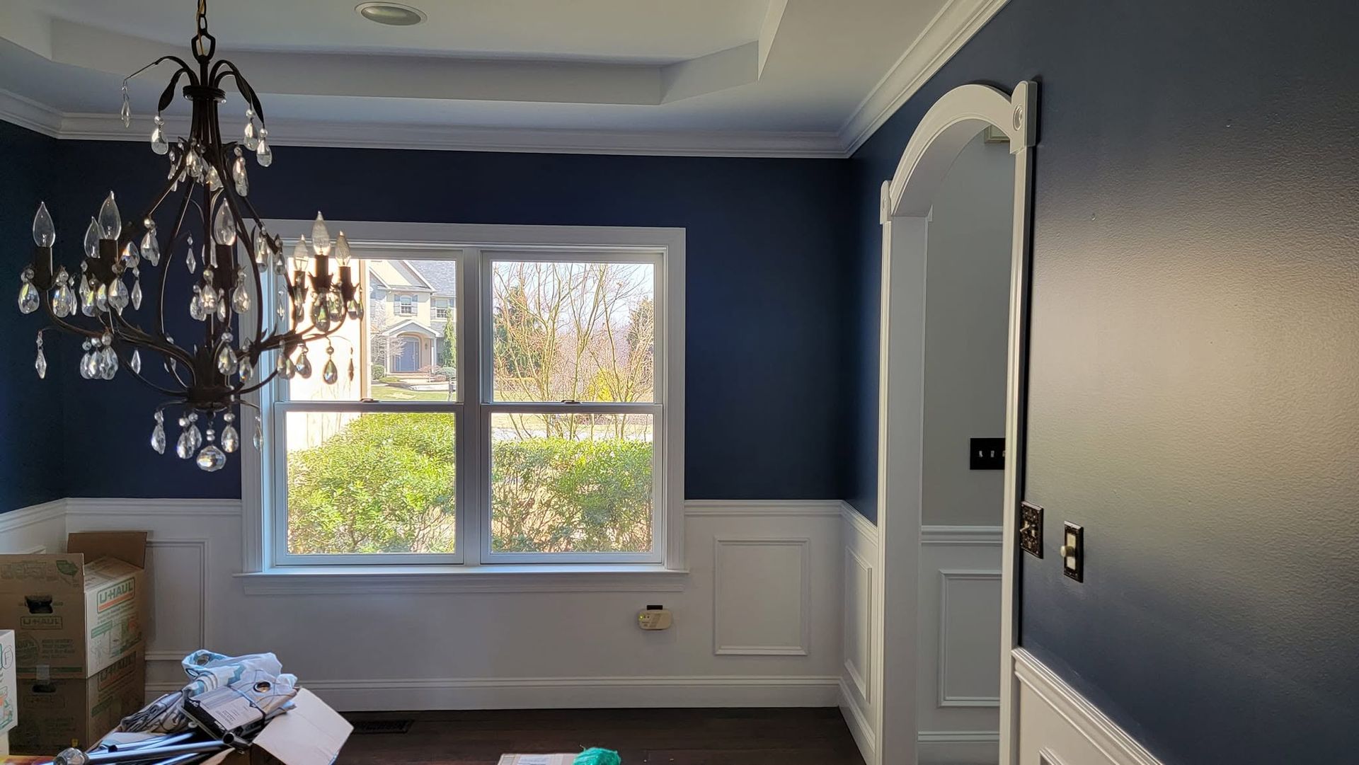 Empty dining room with dark blue walls, white trim, a chandelier, and a large window overlooking greenery.