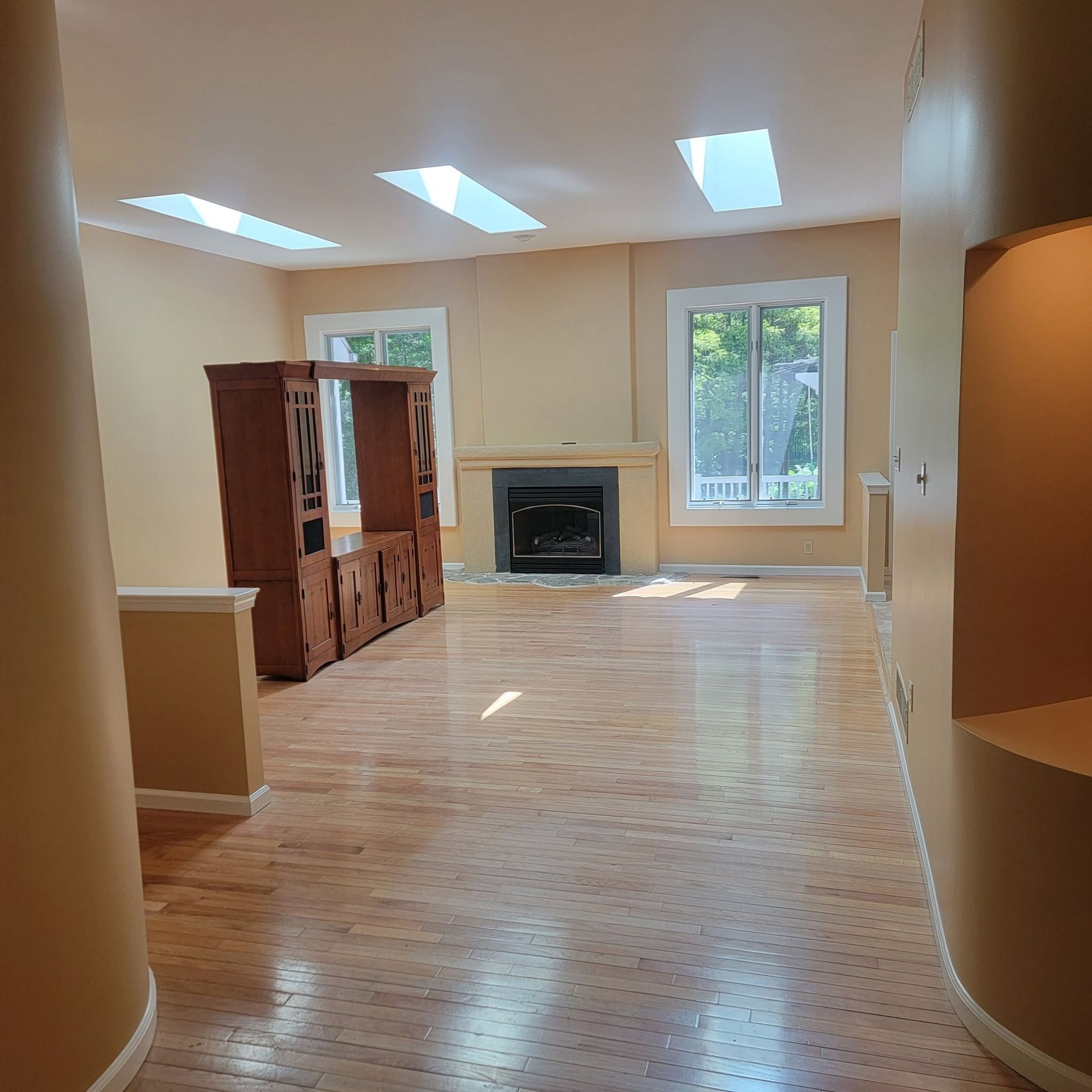 Empty living room with hardwood floors, skylights, fireplace, windows, and a wooden cabinet.