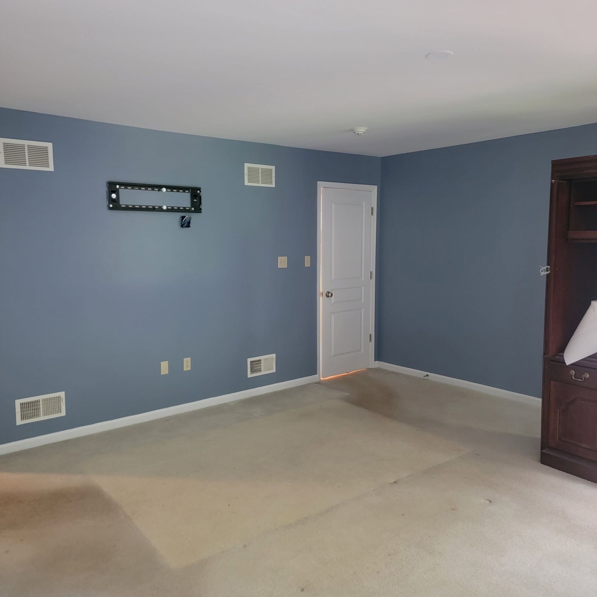 Empty blue bedroom with white door, beige carpet, wall vents, and a dark wooden dresser on the right.