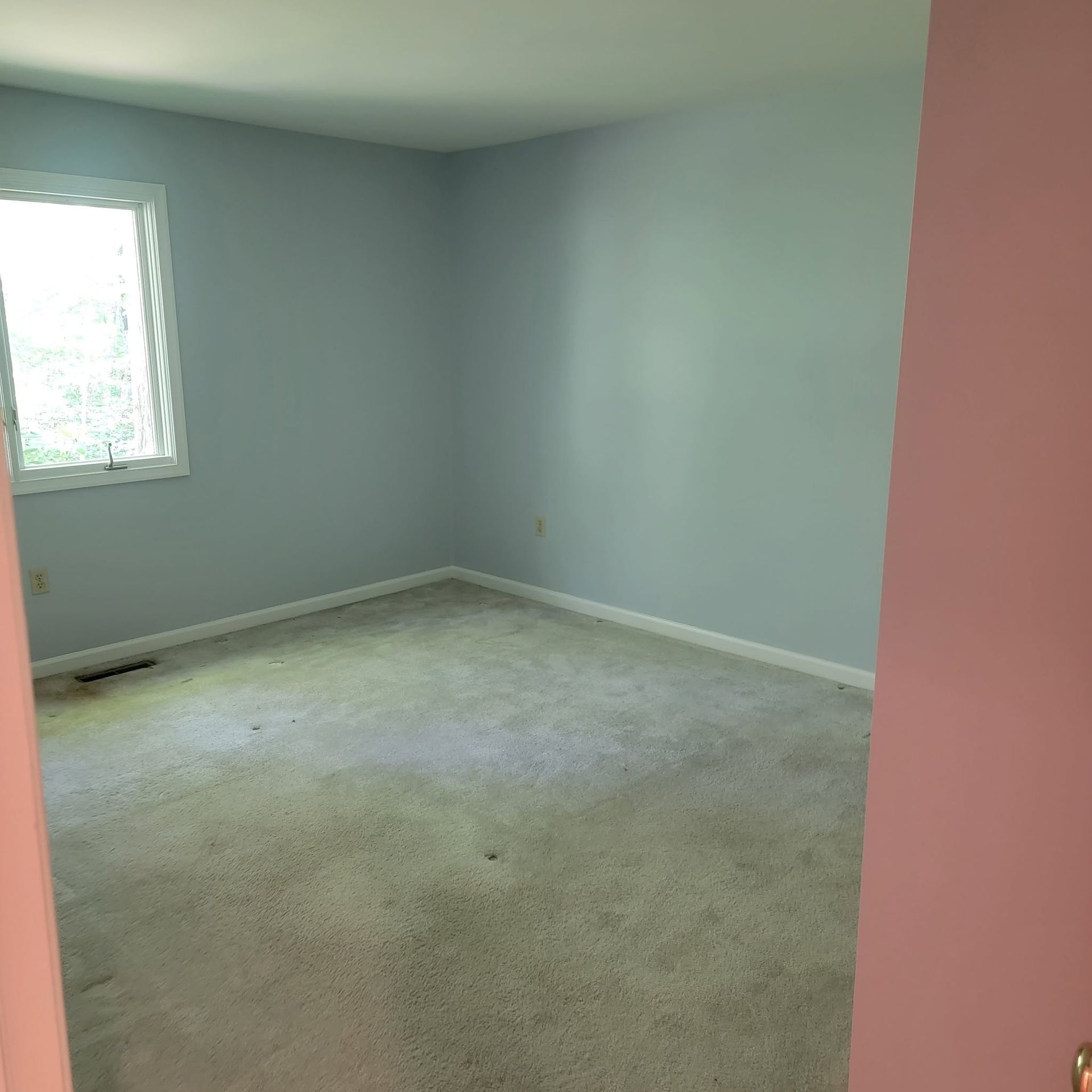 Empty light blue bedroom with a white-trimmed window and worn beige carpet