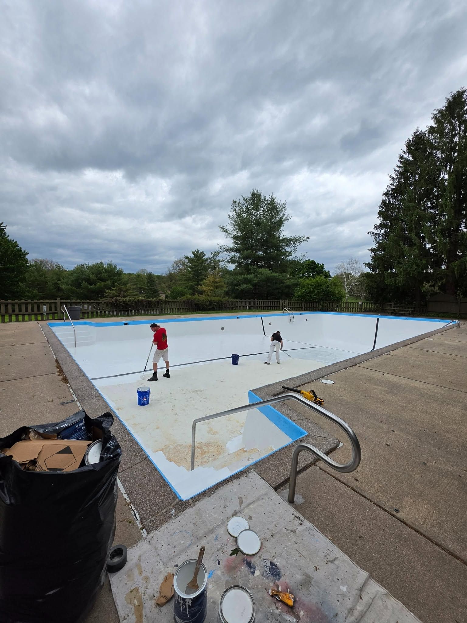 Empty outdoor swimming pool being painted, with buckets and tools on the deck under cloudy skies