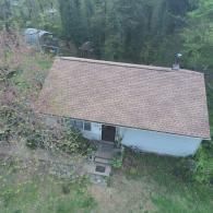 Overhead view of a small, light blue house with a brown roof and overgrown vegetation surrounding it.