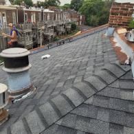 Roofer on a gray shingle roof, working with painting tools, against a city backdrop.