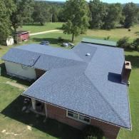 An aerial view of a brick house with a newly installed blue shingle roof in a grassy rural setting.