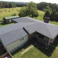Aerial view of a home with a dark gray roof, a porch, and a chimney, set in a rural landscape with trees and fields.