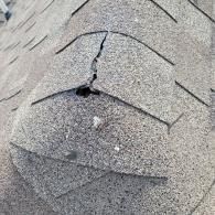 Close-up of damaged asphalt shingles on a roof with a large crack and hole.