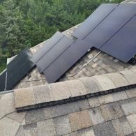 Solar panels of varying sizes installed on a gray-shingled roof against a backdrop of green trees.