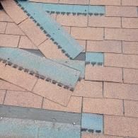 Damaged roof with detached brown shingles, showing blue underlayment.