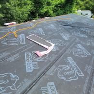 Black roofing underlayment on a roof with a roll of tape and a power cord. Trees and an RV are in the background.