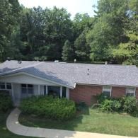 Brick house with gray roof, green bushes, and a curved walkway. Trees in the background.