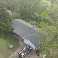 Overhead view of a gray-roofed house surrounded by trees, with debris near the house.