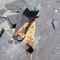 Damaged roof with missing shingles, exposing rotted wood and a hole, next to a pipe.