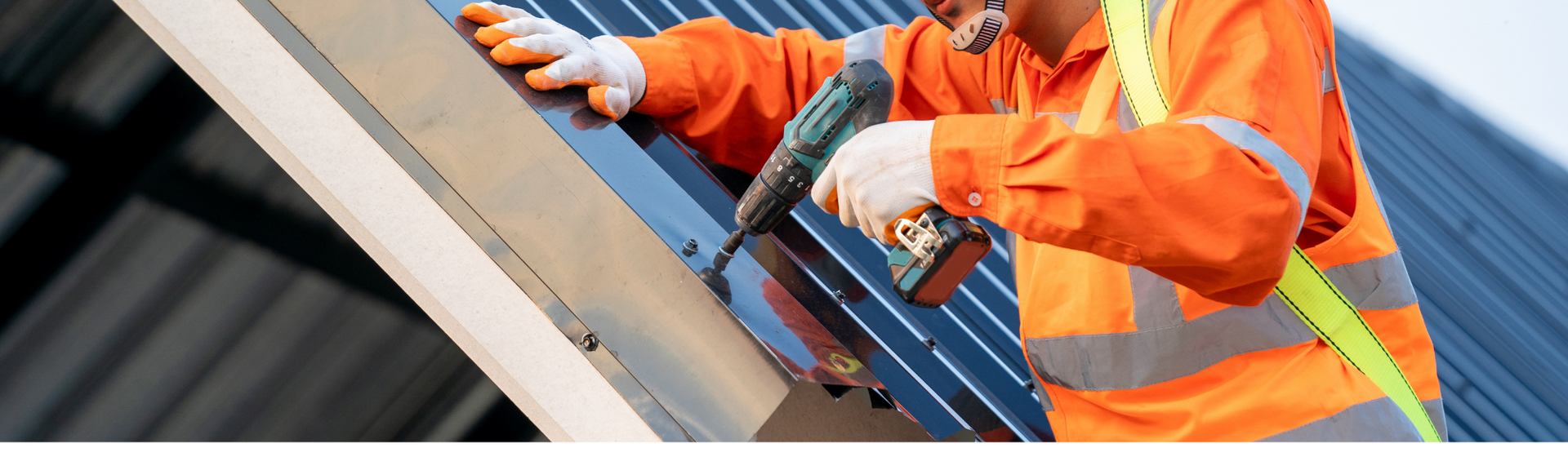 A person in orange workwear installs solar panels on a roof, using a power tool.
