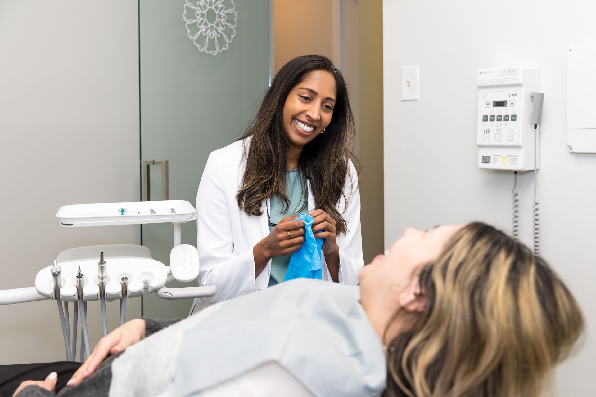 Dentist preparing for patient. Dentist in white coat smiles holding gloves; patient reclines in dental chair.