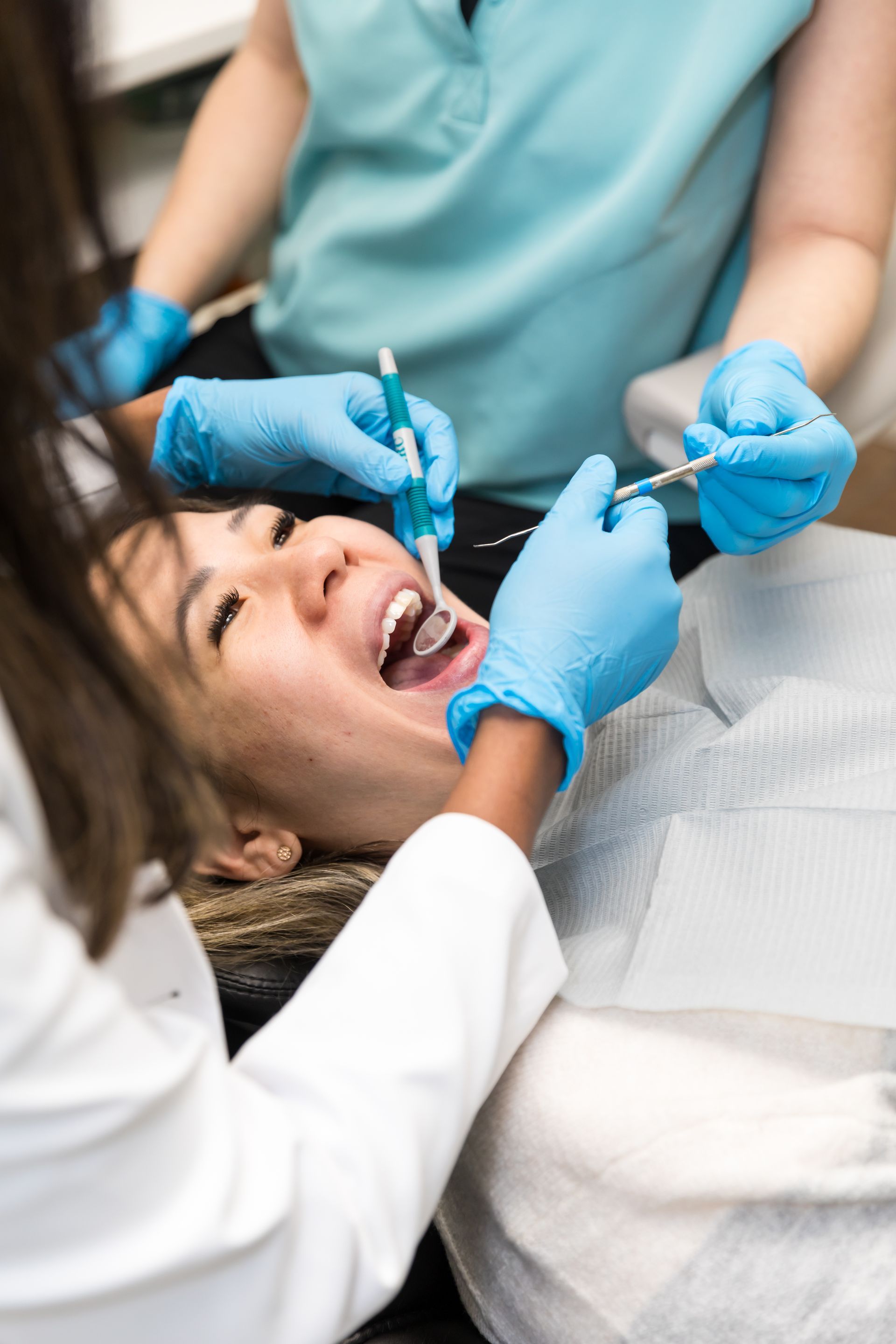 Woman at dentist, mouth open, receiving dental care by two people wearing blue gloves.