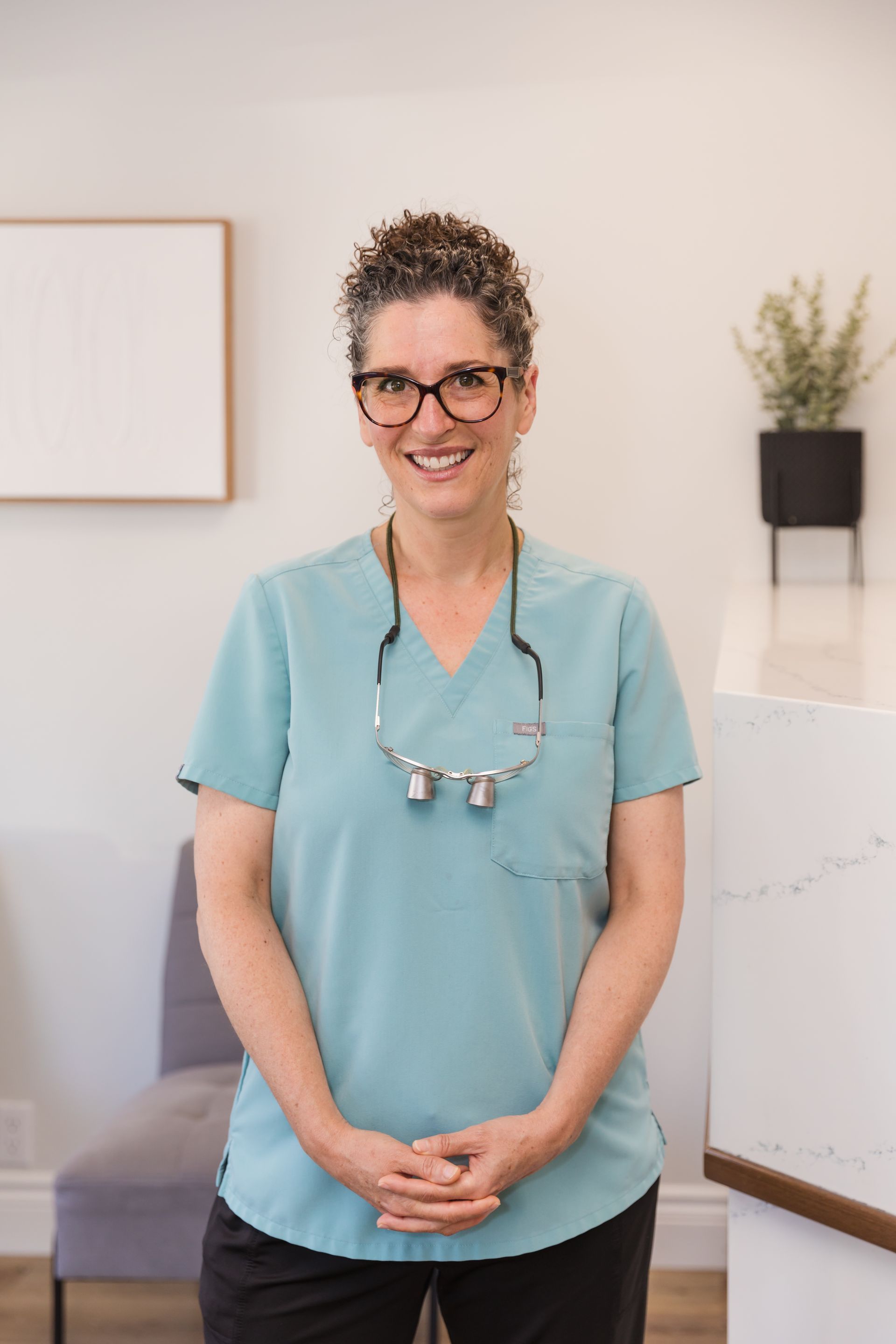 Woman in teal scrubs smiles, wearing glasses and loupes, in a dental office.