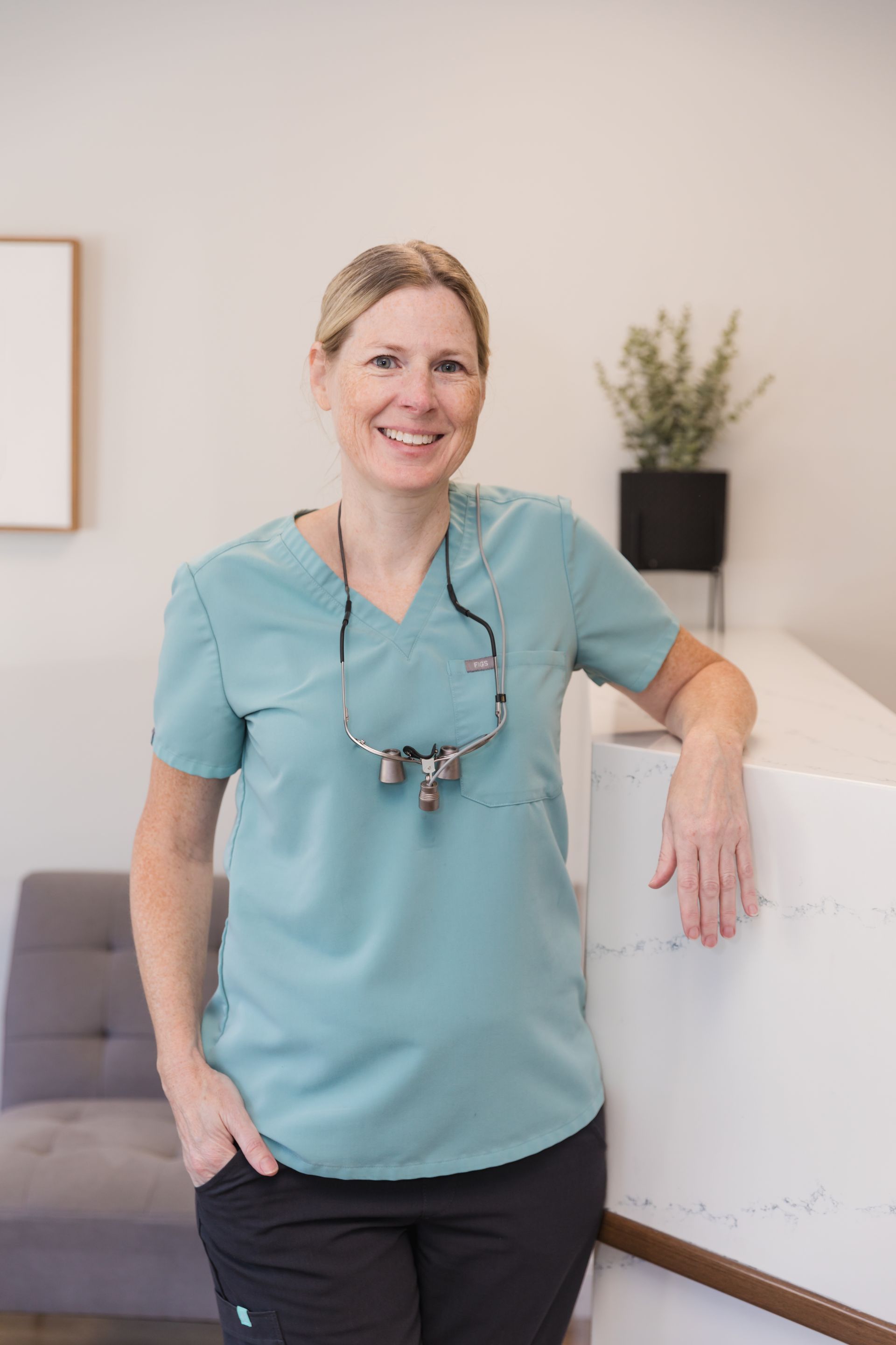 Dentist smiling, leaning on a white counter in a modern office, wearing teal scrubs.