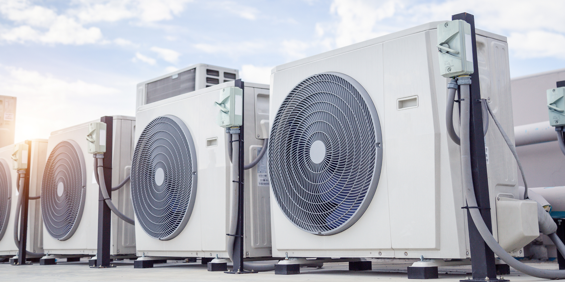 Air conditioning units on a rooftop, lined up. Bright sky and sunlight in the background.
