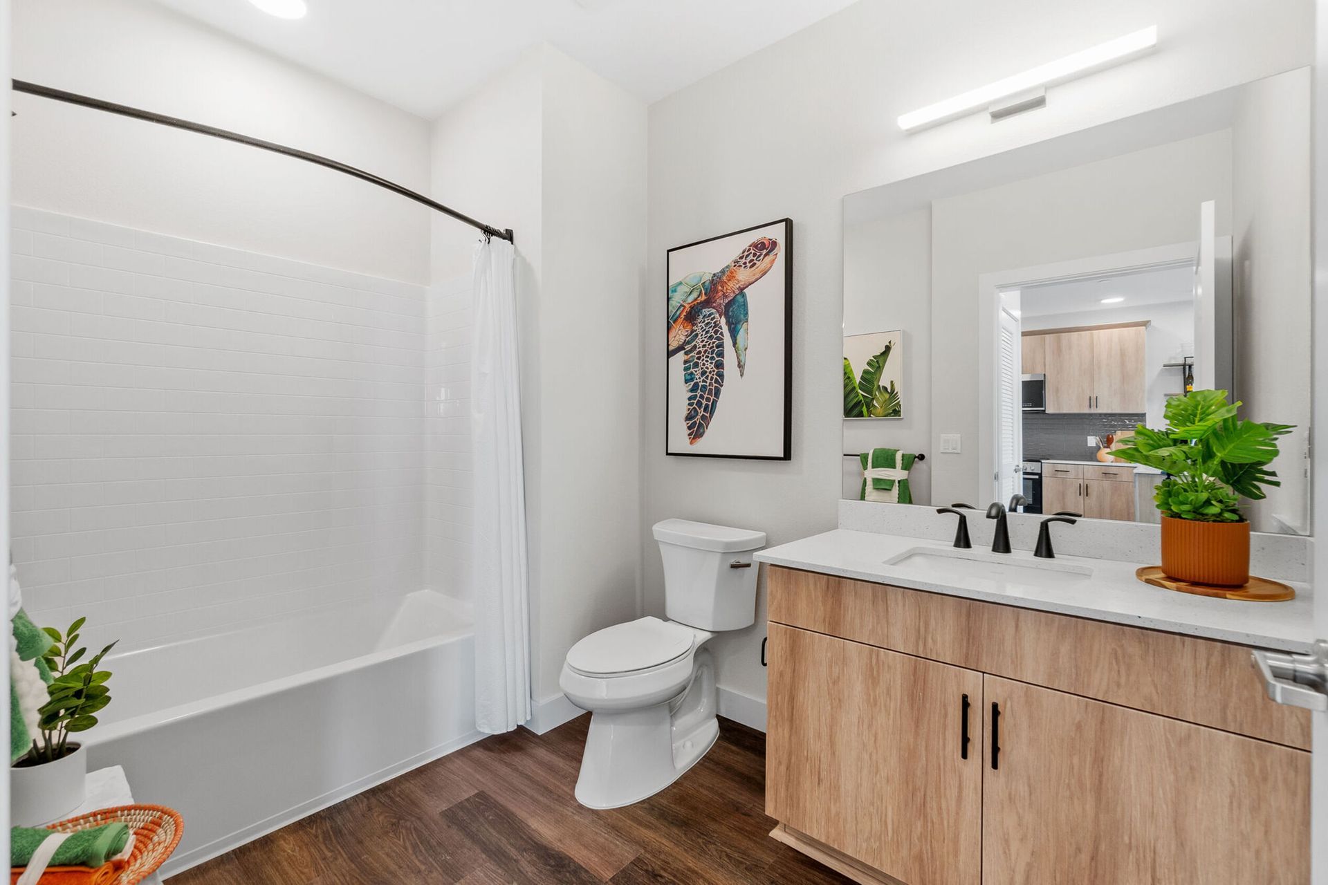 Bathroom with wood vanity, white toilet, bathtub, art, and plant. Dark wood floor.
