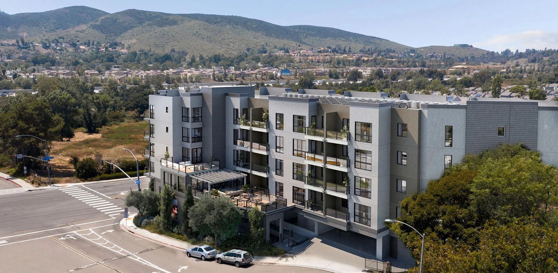 Multi-story building with gray exterior and balconies, trees, cars on a road, and hills in the background.
