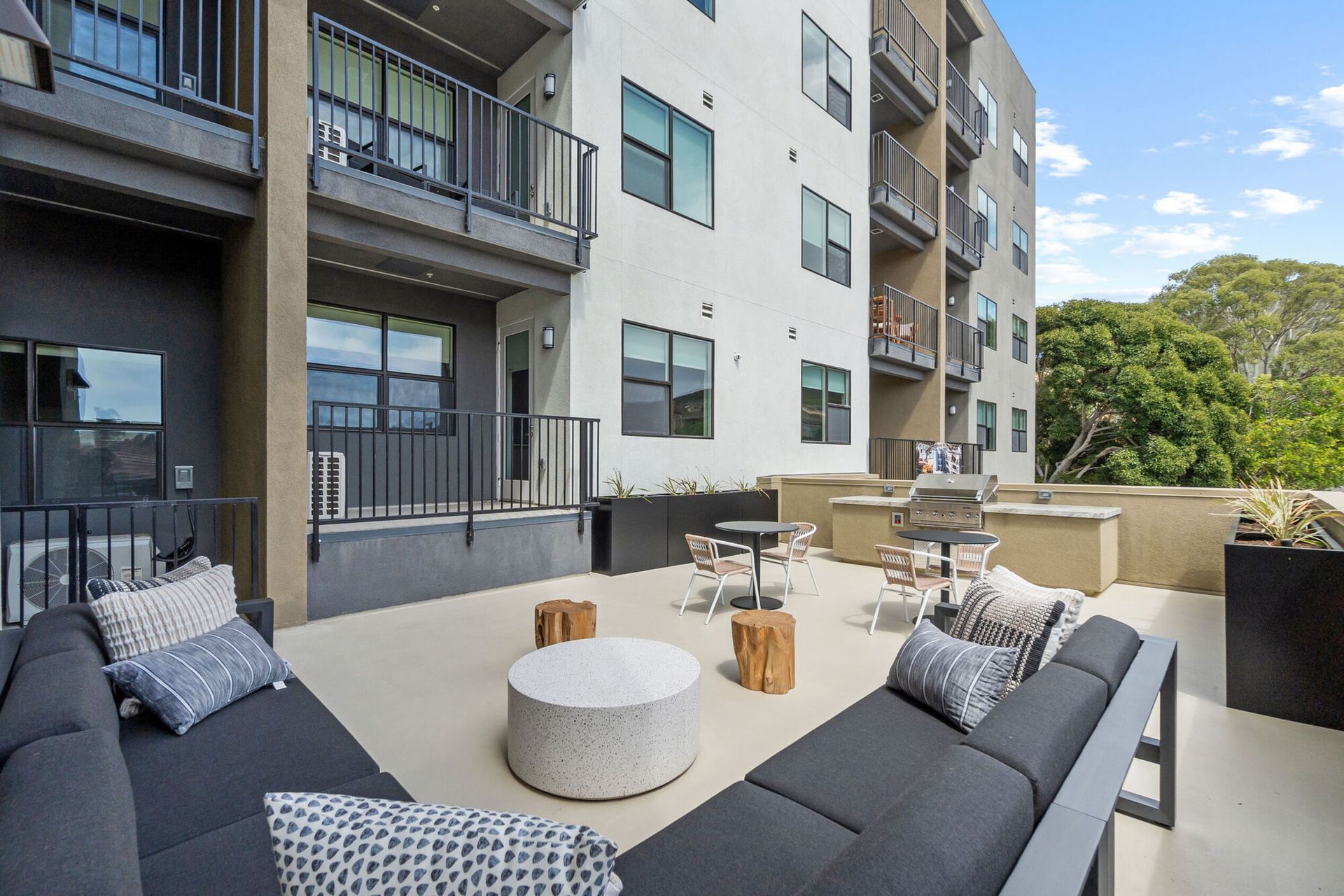 Outdoor patio with gray couches, small tables, and grill. Apartment building in background.