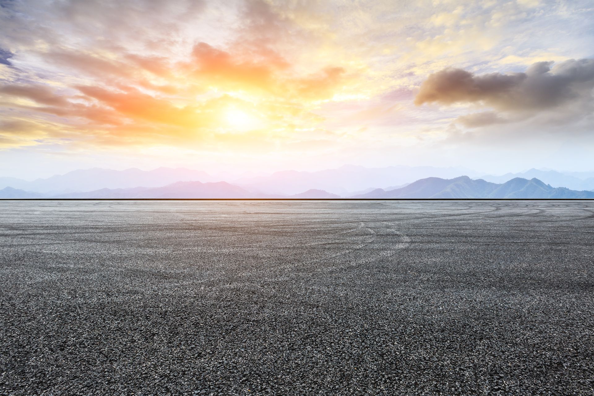 Person using a tool to smooth hot asphalt on a road; steam rising, orange machine in the background.