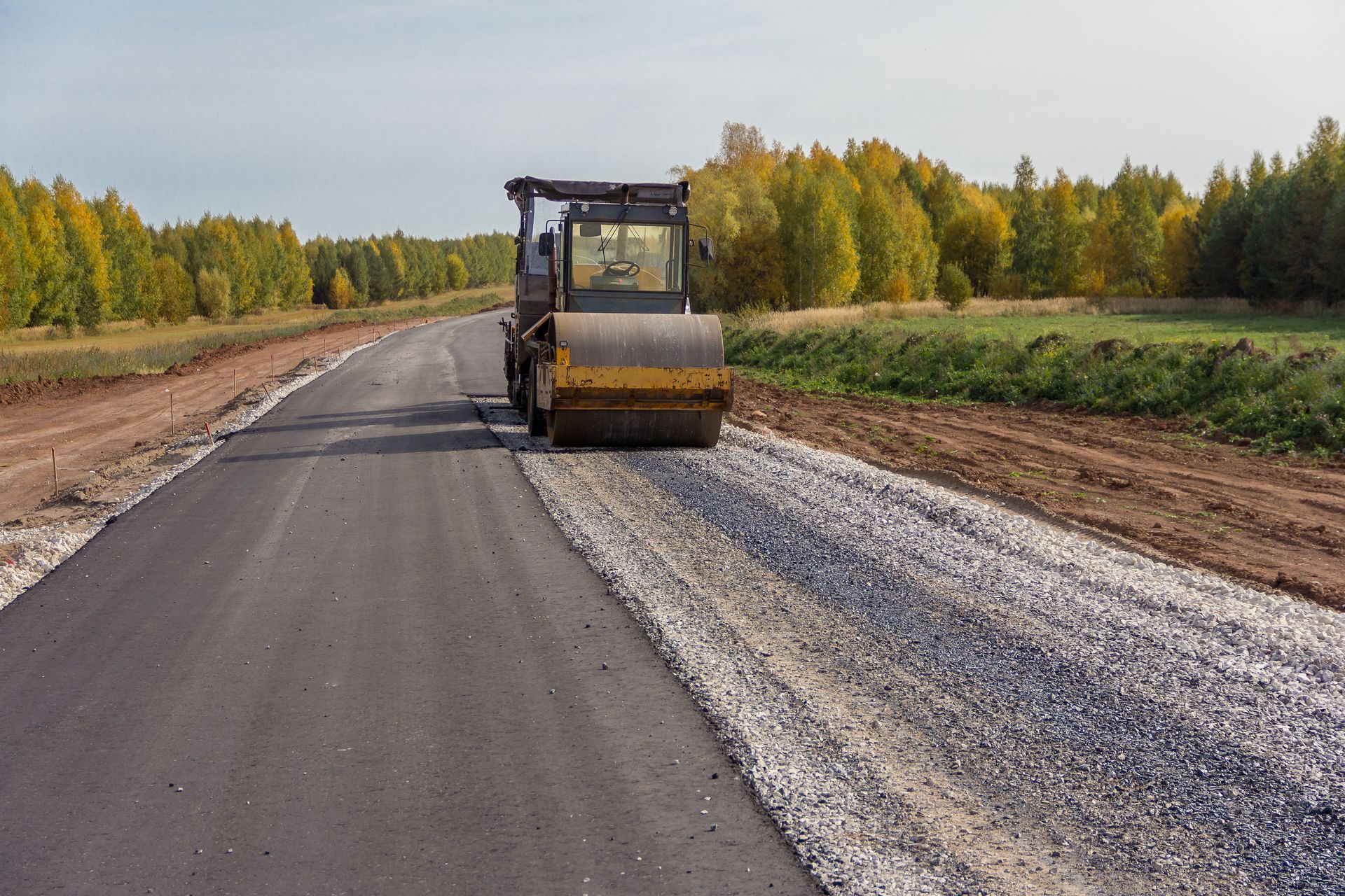 Road roller compacting asphalt on a newly paved road. Gravel and trees are visible in the background.