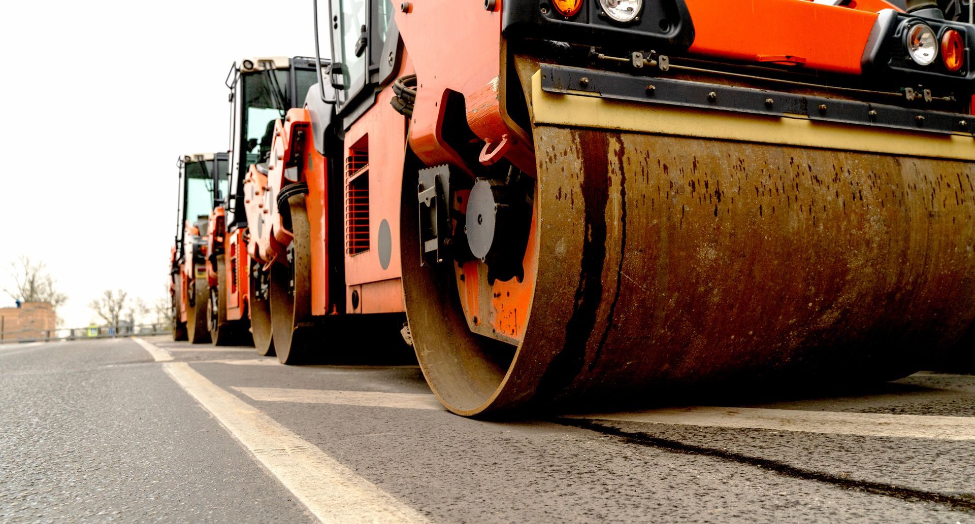 Orange road roller compacting asphalt on a road with other machinery in the background.