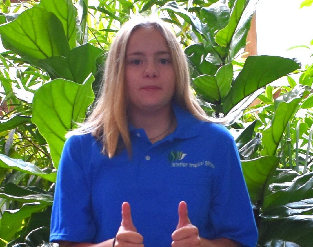 Person in blue polo shirt with thumbs up, standing in front of lush green foliage.