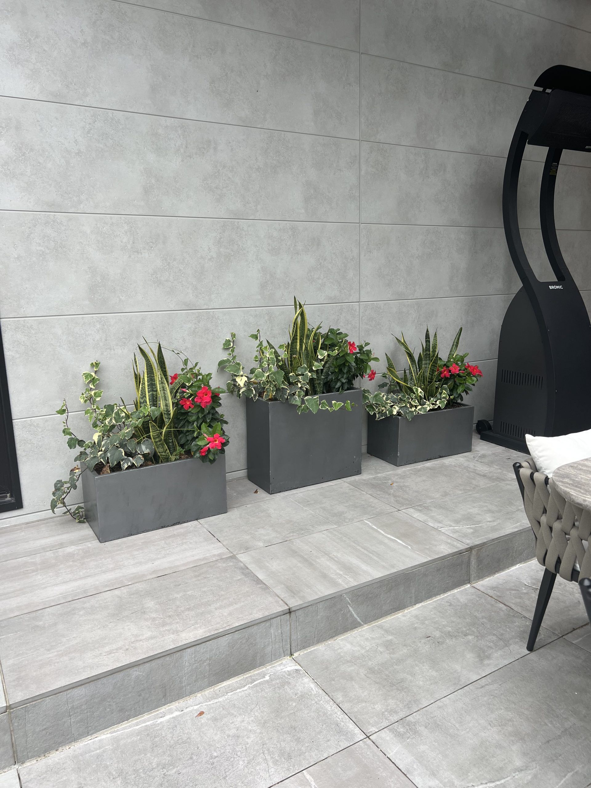 Three rectangular planters with flowers on a gray patio next to a heater.